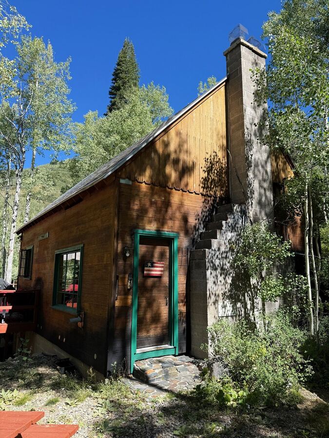 The front of the cabin with a green door, stone chimney and a small flag beside the doorway